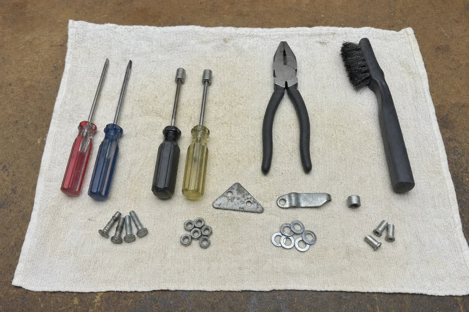 Four drivers, pliers and a wire brush on a shop towel with a bracket, link, spacer and sorted bolts and washers