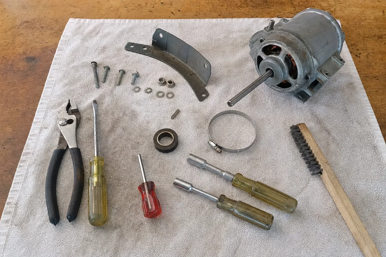 Small electric motor with its shaft bare, a curved mounting bracket, hose clamp, bushing and bolts beside hand tools